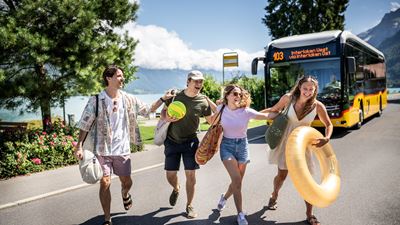 Eine Gruppe Jugendlicher mit Badeutensilien überquert eine Strasse am Thunersee vor einem Postauto.
