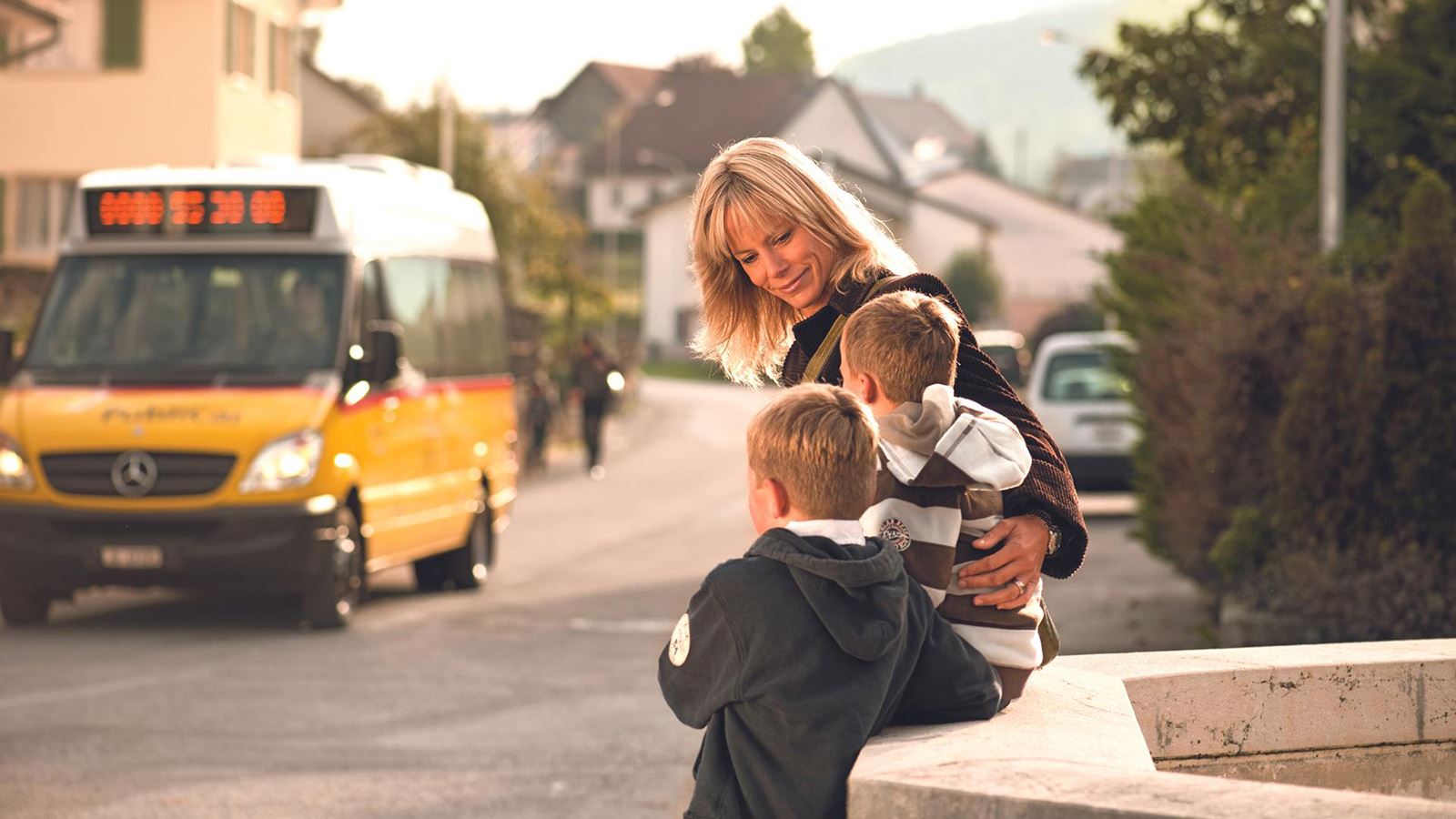 Une mère attend avec deux enfants à l’arrêt CarPostal. Un car postal est visible en arrière-plan.