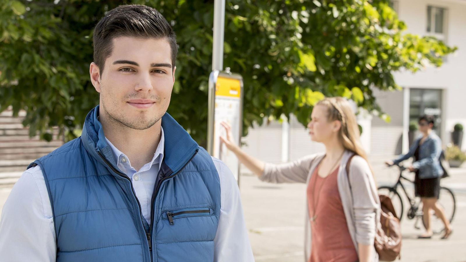 Un uomo si trova a una fermata dell’autopostale. Sullo sfondo qualcuno guarda l’orario.