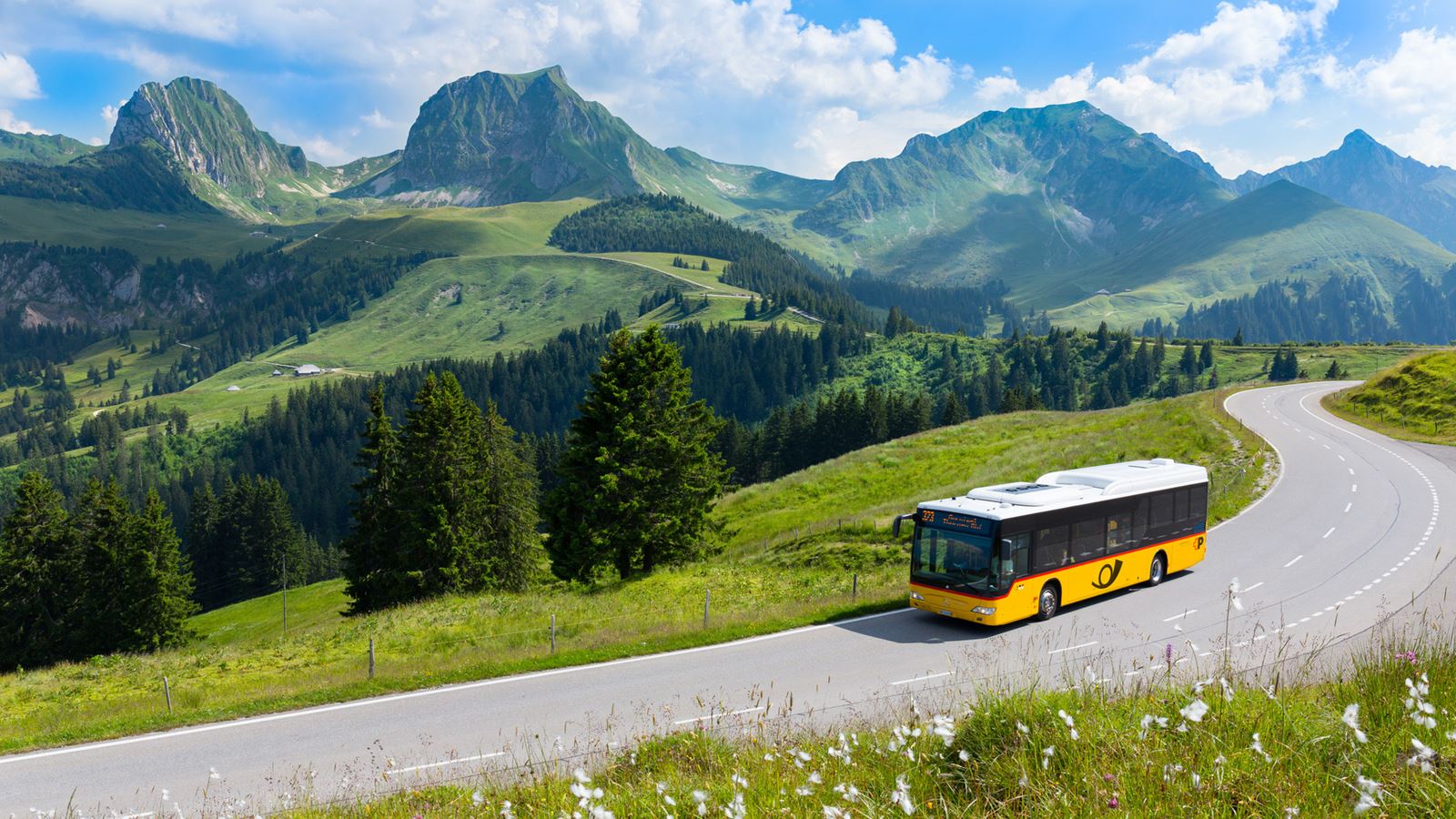 Postbus driving along a road. A green meadow in the foreground and the Gantrisch mountain range in the background.