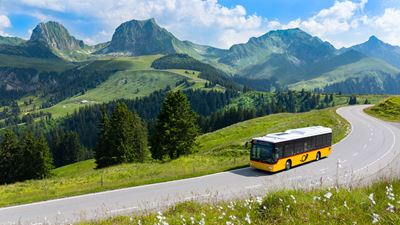 Postbus driving along a road. A green meadow in the foreground and the Gantrisch mountain range in the background.
