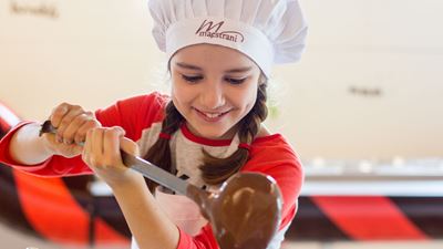 Une petite fille portant une toque Maestrani verse du chocolat fondu dans une casserole à l’aide l’une louche. © Maestrani Chocolats Suisses SA