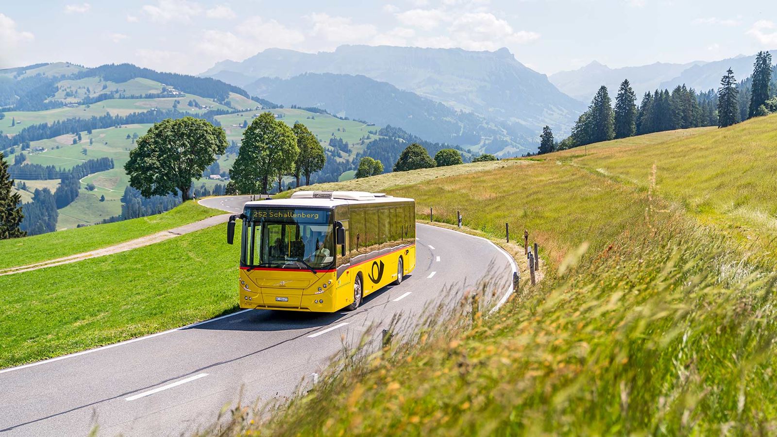 CarPostal circule sur la route dans un environnement rural. Au fond, le paysage de collines de l’Entlebuch avec vue sur la Schrattenfluh.