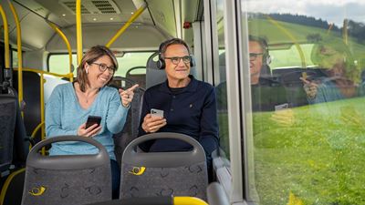 A woman and a man listening to the Umweg am Albis audio experience in a Postbus. They’re wearing headphones and looking out the window.