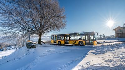 A yellow Postbus travels through the snowy landscape over the Albis Pass in the sunshine.