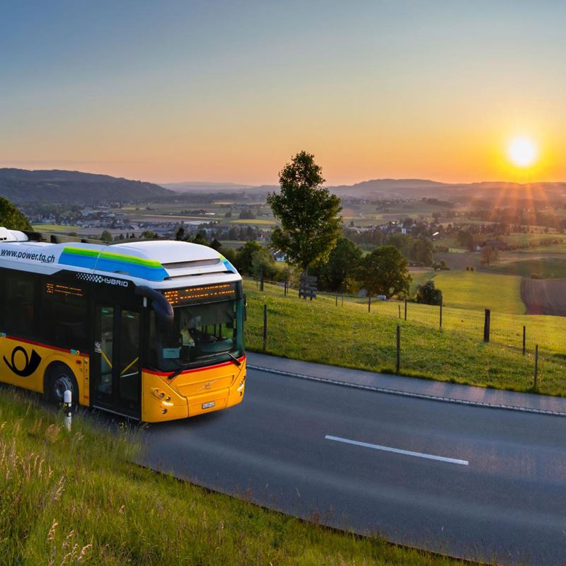 Ein gelbes Postauto fährt auf einer Landstrasse, umgeben von grünen Feldern und sanften Hügeln, während die Sonne am Horizont untergeht. Die friedliche Landschaft und das warme Licht der untergehenden Sonne schaffen eine ruhige und idyllische Atmosphäre.,Ein gelbes Postauto fährt auf einer Landstrasse, umgeben von grünen Feldern und sanften Hügeln, während die Sonne am Horizont untergeht. Die friedliche Landschaft und das warme Licht der untergehenden Sonne schaffen eine ruhige und idyllische Atmosphäre.