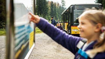 A child looks at the timetable at a Postbus stop and there is a Postbus in the background.