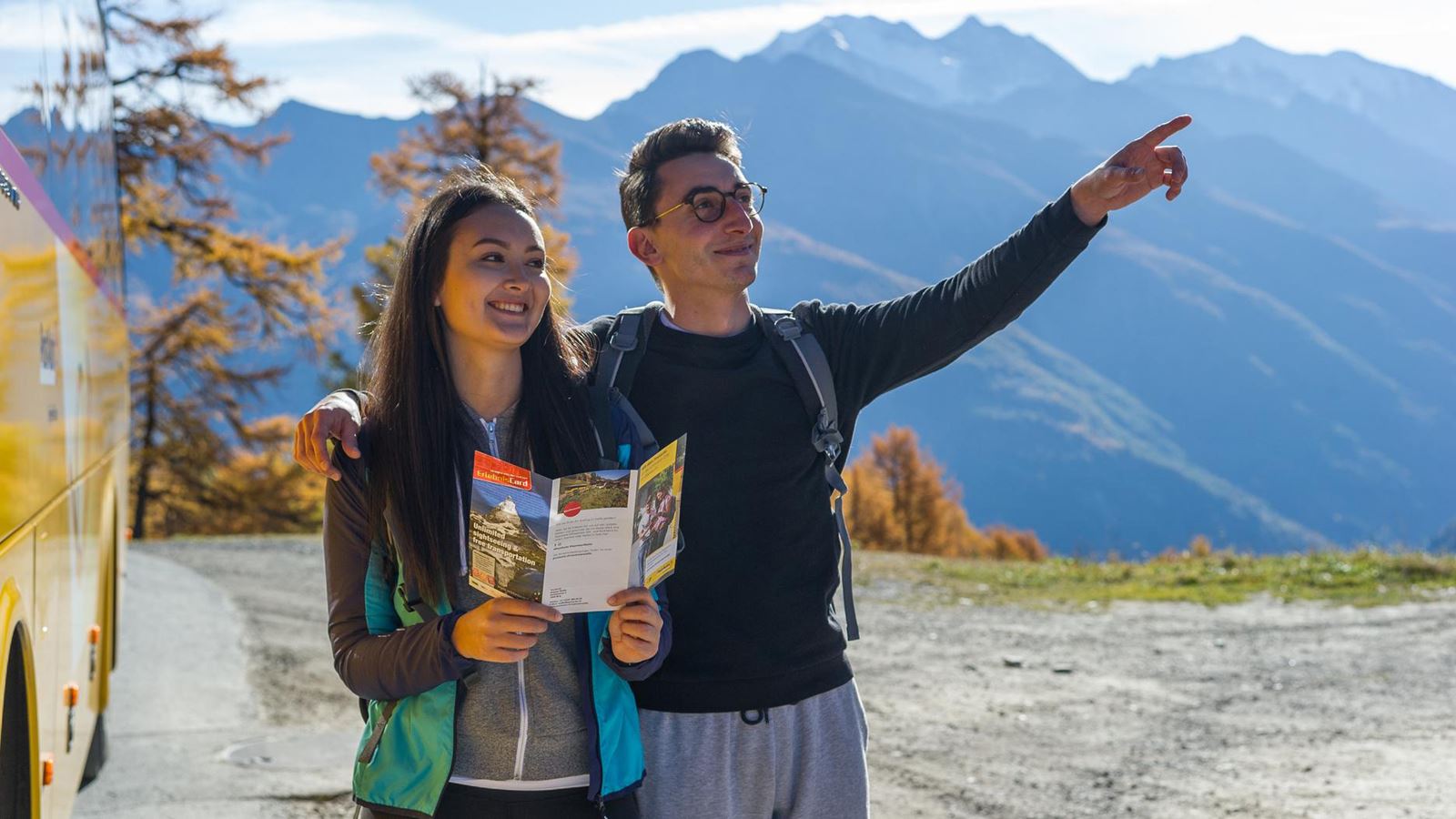 A man and a woman stand in front of a PostBus and plan their way.