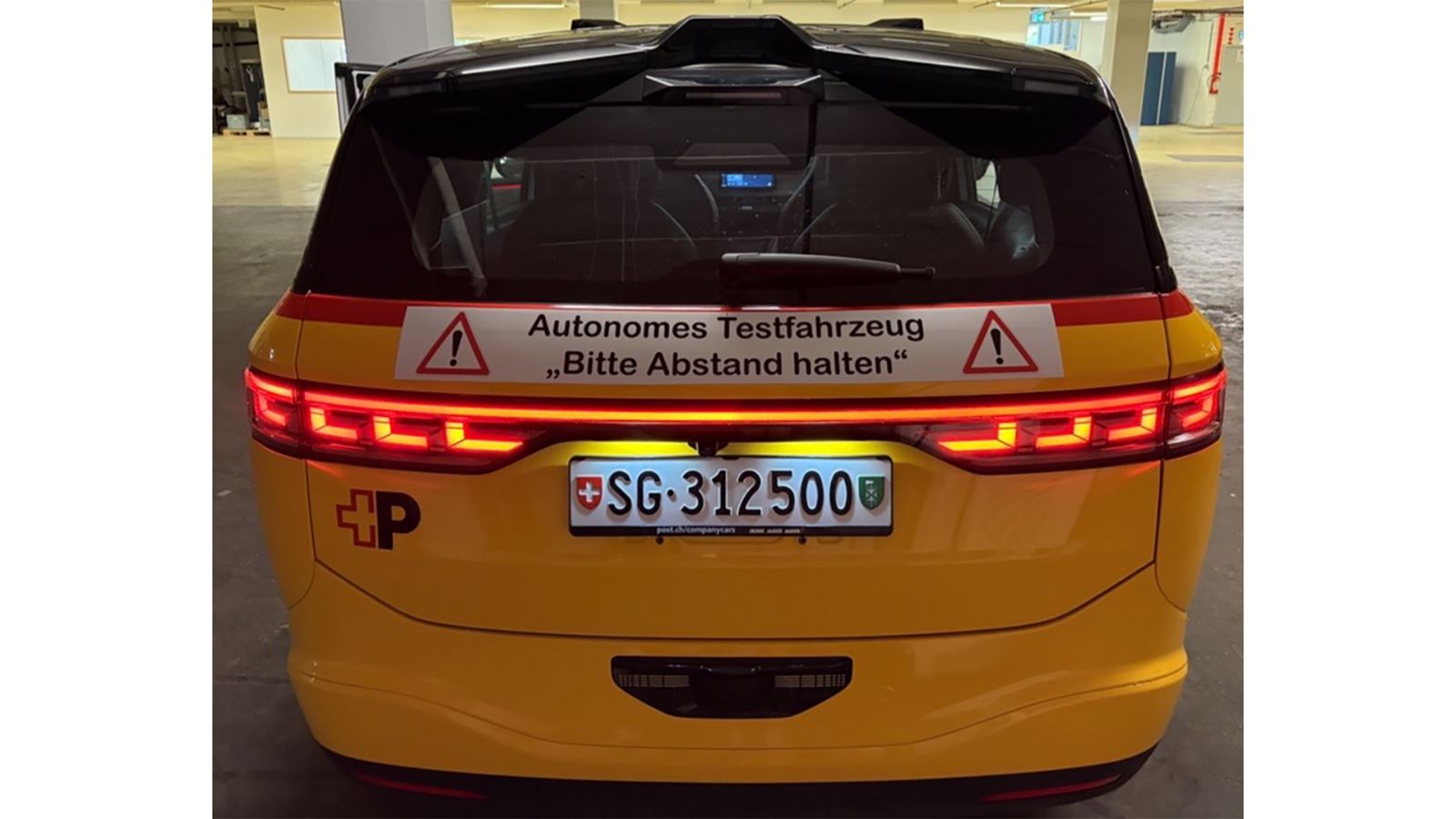 Un veicolo AmiGo giallo visto dalla parte posteriore. Sopra la targa è presente un manifesto con la scritta «Veicolo di prova autonomo, mantenere la distanza!». 