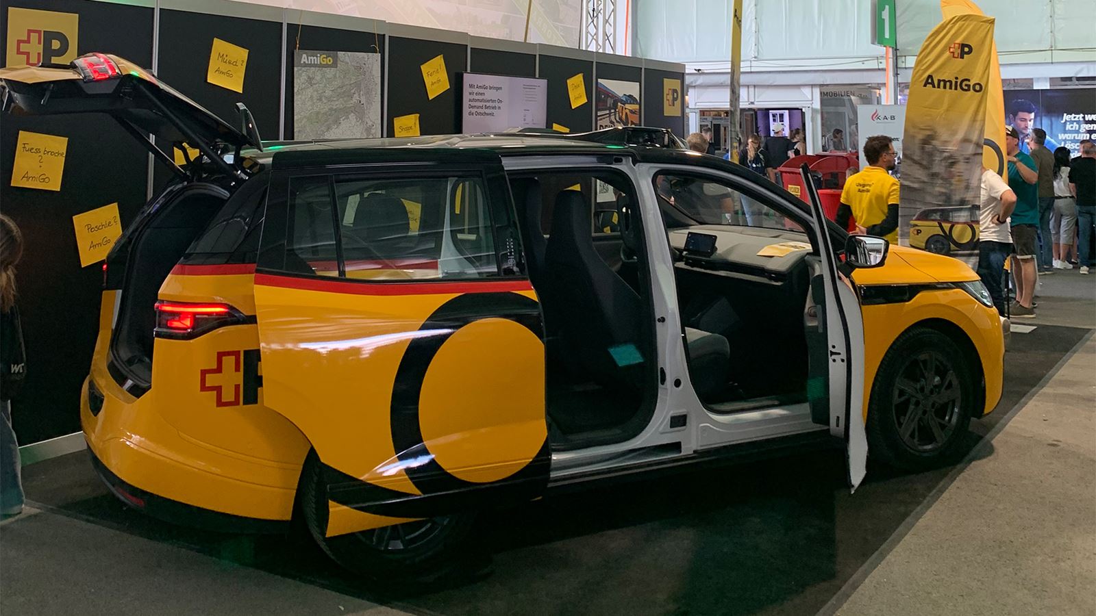 Dans une halle du salon, un véhicule AmiGo jaune se trouve portes ouvertes sur le stand. En arrière-plan, il y a des panneaux d’information ainsi que des visiteuses et des visiteurs.