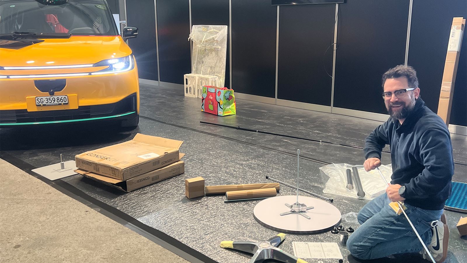 Montage d’un stand: le véhicule AmiGo jaune est prêt, une personne monte le stand sur la surface.