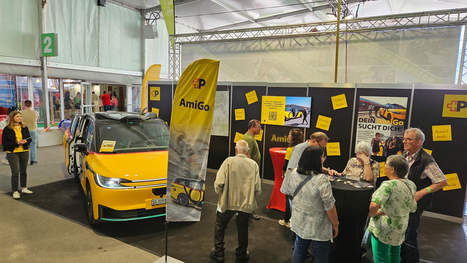 Messestand «AmiGo» mit gelbem AmiGo-Fahrzeug; mehrere Besuchende informieren sich an Tafeln und Bildschirmen.
