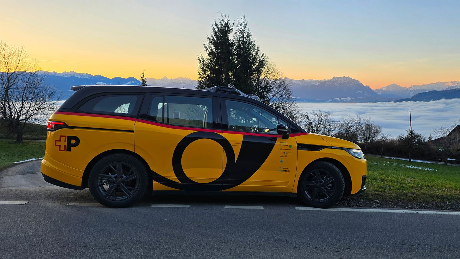 Gelbes, autonomes AmiGo Fahrzeug steht am Straßenrand vor Alpenpanorama mit Nebelmeer im Hintergrund in der Schweiz.