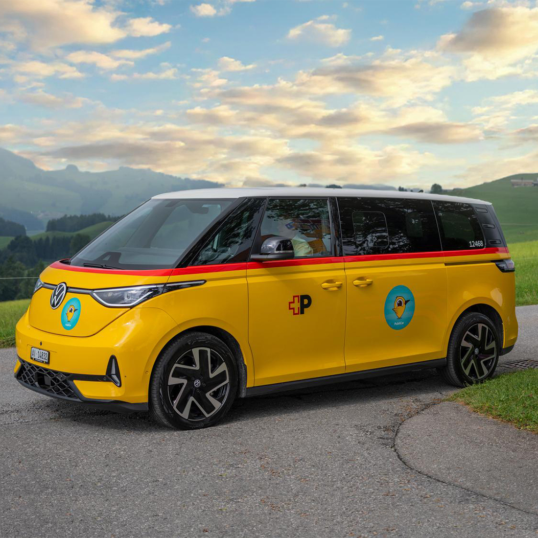 Bus VW électrique au design jaune CarPostal avec bande rouge et logo du cor postal, utilisé pour le service à la demande PubliCar à Appenzell. Le véhicule est stationné sur une route de campagne dans un paysage vallonné pittoresque de Suisse, à côté d’un panneau indicateur pour les chemins de randonnée pédestre et cyclable