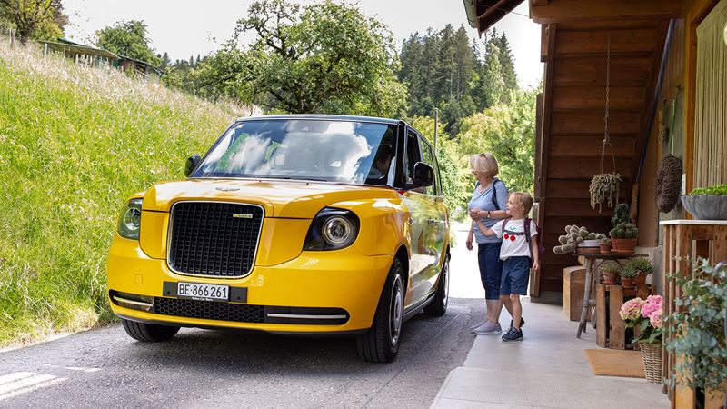 Eine Frau und eine Kind welches rechts vor dem Postauto London Taxi stehen. Das Postauto London Taxi wird frontal gezeigt.