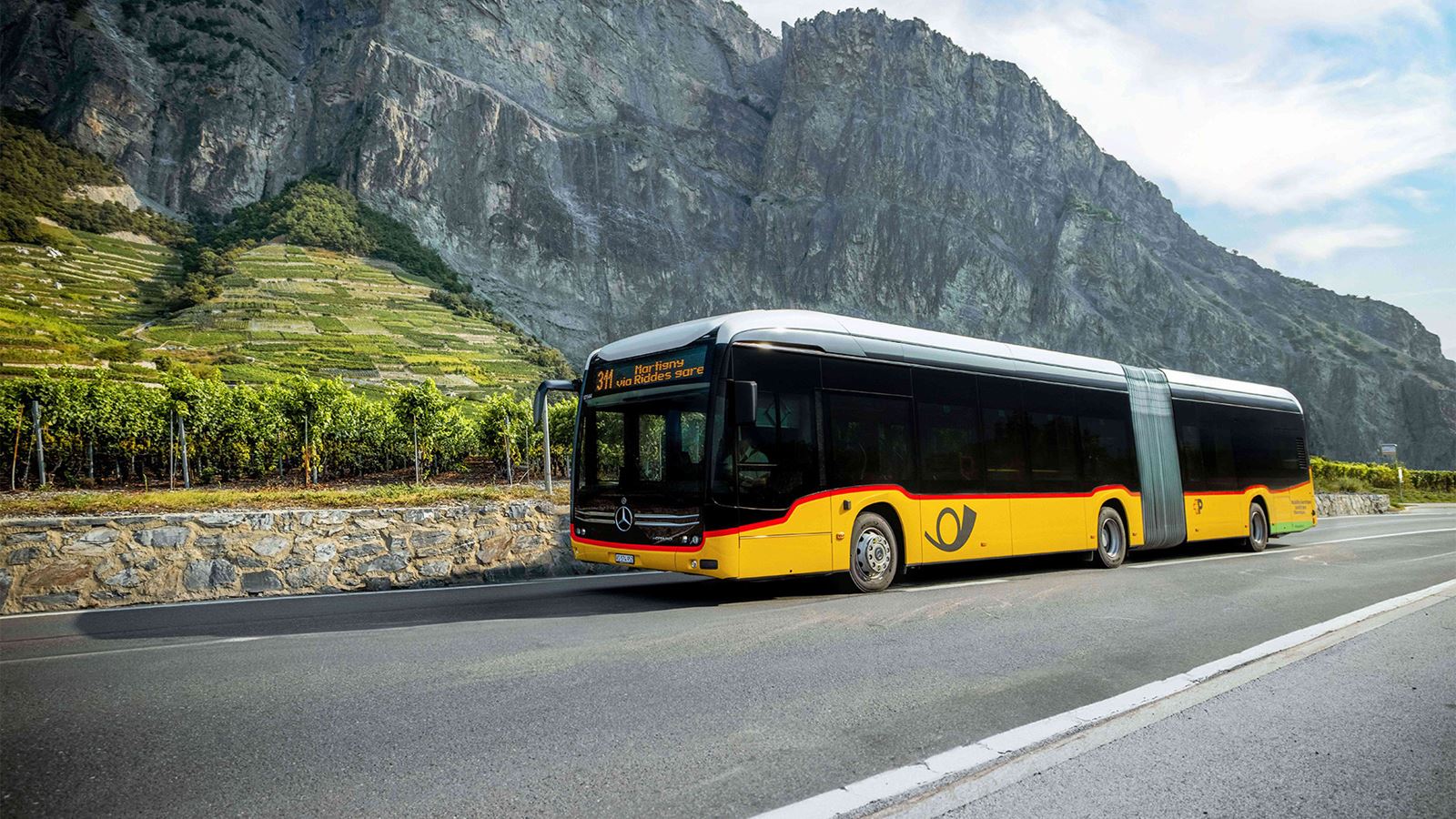 Un car postal articulé électrique sur une route, devant une montagne, au pied des vignes